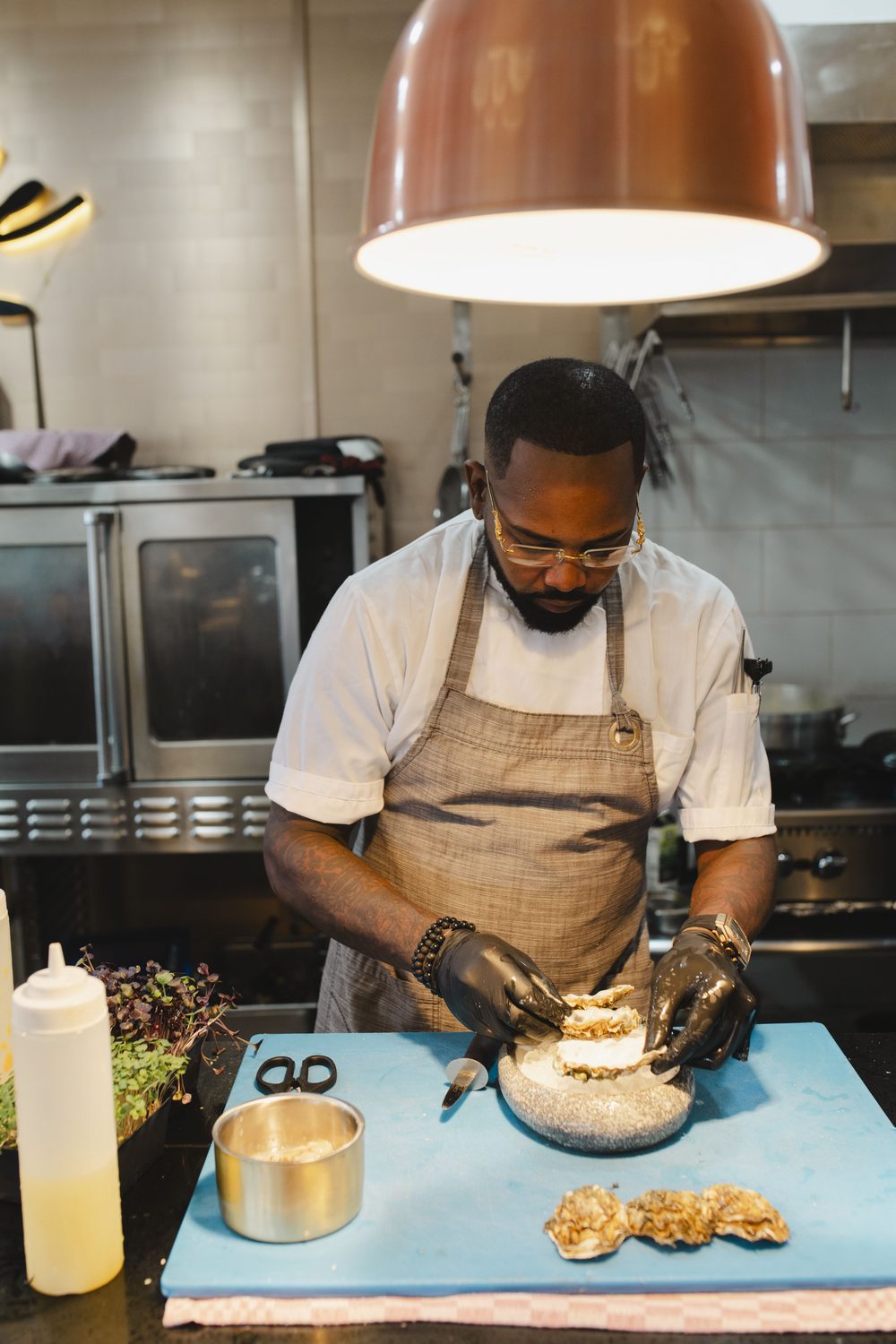 Chef Moisés Carvajal shucking oysters at the pass, under a copper lamp