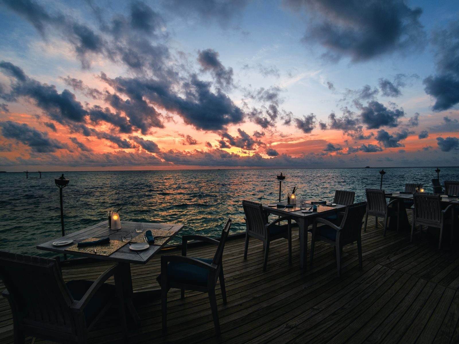 An open-air deck over the water at dusk, wooden tables laid with lit candle lanterns, the sea and a streaked orange sky beyond