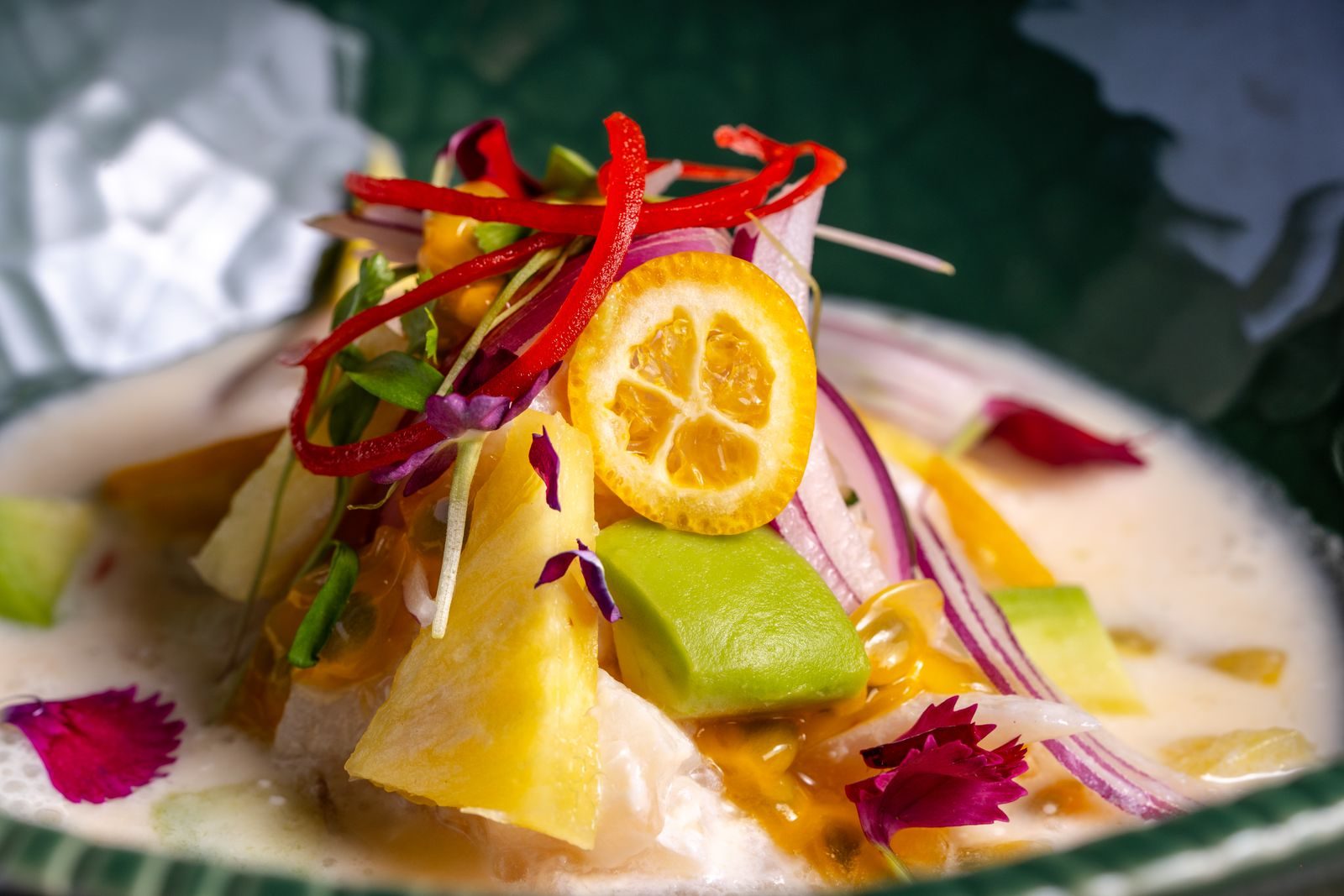 Slices of raw white fish in a milky leche de tigre with red onion, chilli, avocado and micro greens on a dark green plate
