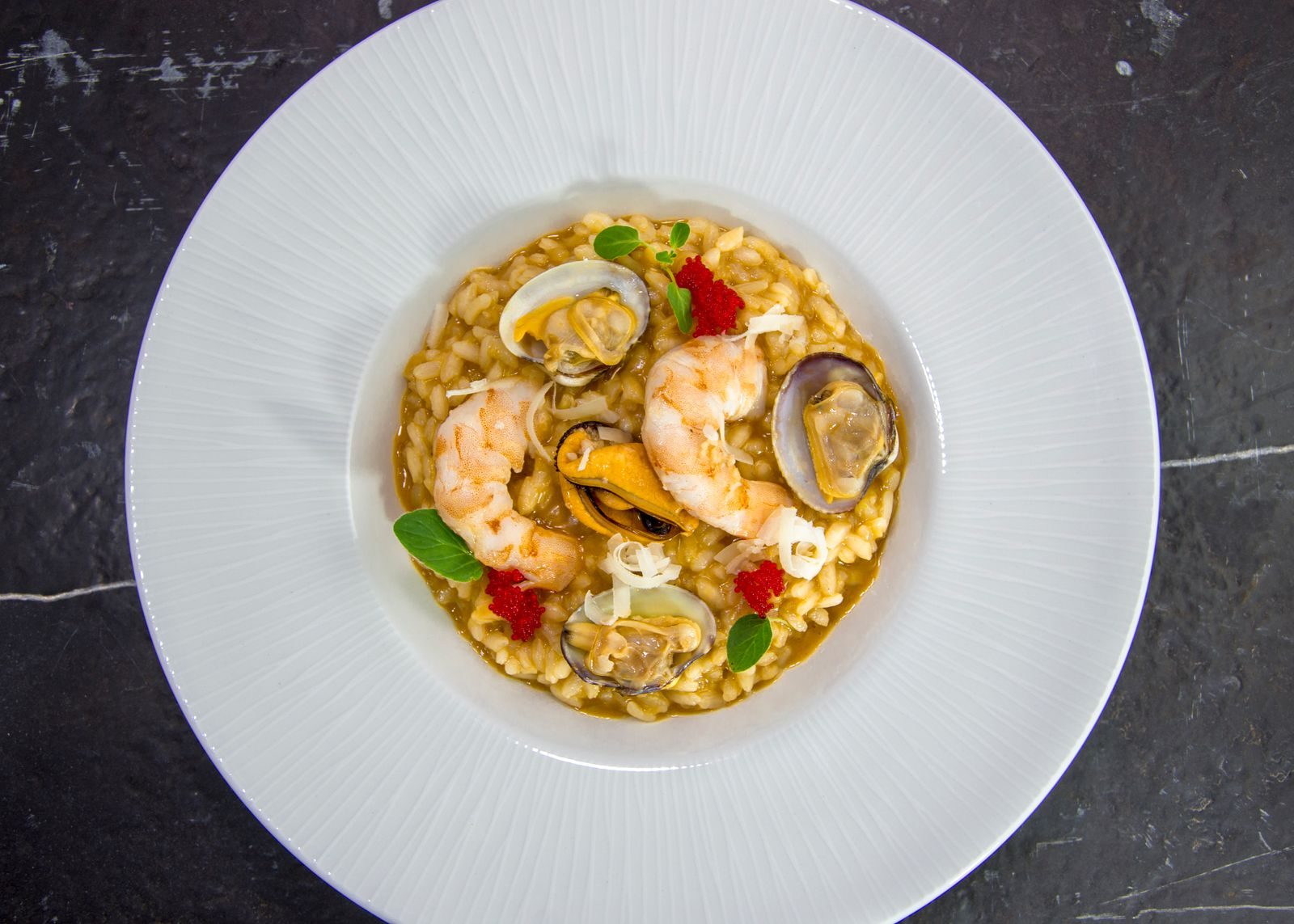 A saffron-coloured seafood risotto with prawns, open clams and a mussel, on a wide white plate over dark stone