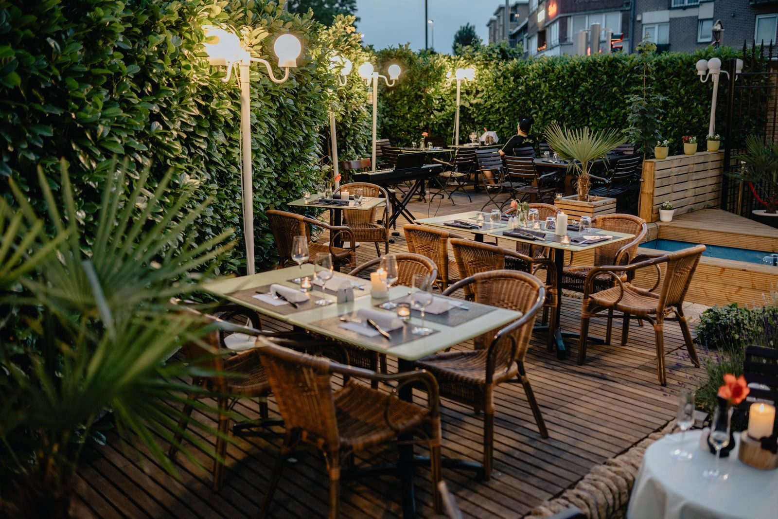 A restaurant terrace at blue hour, several tables laid and candlelit under lit lamp-posts, palm fronds at the edge of frame