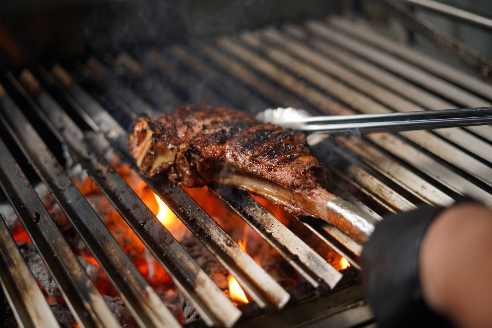 A thick bone-in steak on a heavy steel grill grate with live flame licking up between the bars, a gloved hand and tongs at the edge of frame