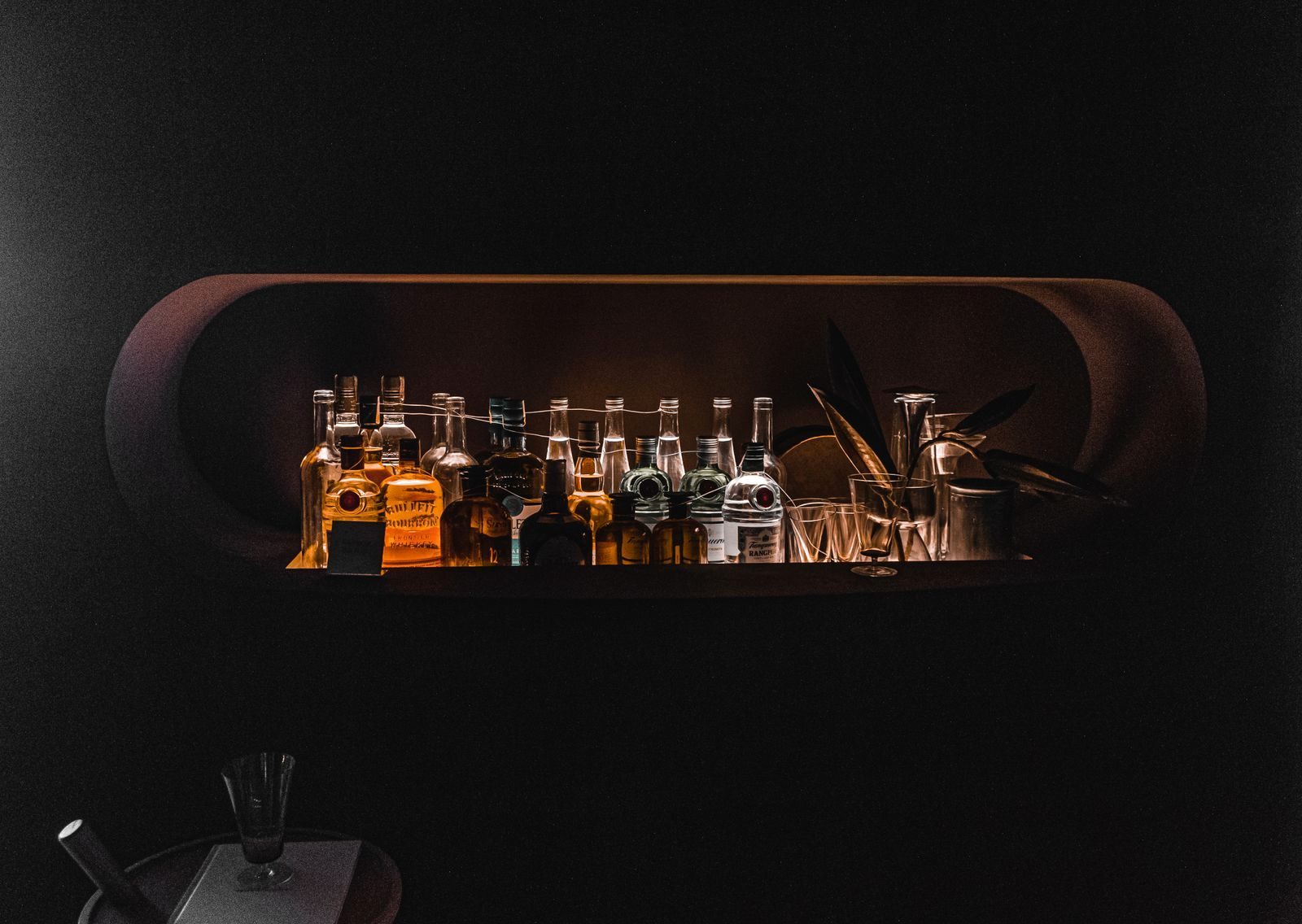 A dark bar wall with a single lit alcove holding a row of bottles and glassware